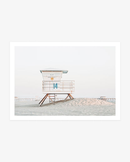 Huntington Beach lifeguard tower wall art featuring Tower 14 on a sandy rise with the pier visible in the distance beneath a pale coastal sky.