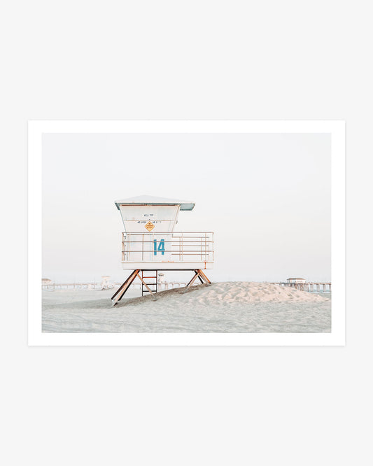 Huntington Beach lifeguard tower wall art featuring Tower 14 on a sandy rise with the pier visible in the distance beneath a pale coastal sky.
