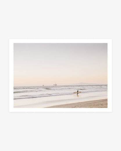 Huntington Beach surfer wall art featuring a lone surfer carrying a board along the shoreline with Catalina Island and offshore platforms in the distance.