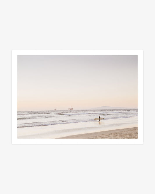 Huntington Beach surfer wall art featuring a lone surfer carrying a board along the shoreline with Catalina Island and offshore platforms in the distance.