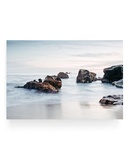 Rocky shoreline at Laguna Beach during sunset with large coastal rocks emerging from soft flowing ocean water, pastel sky and misty long exposure waves creating a calm coastal landscape scene.