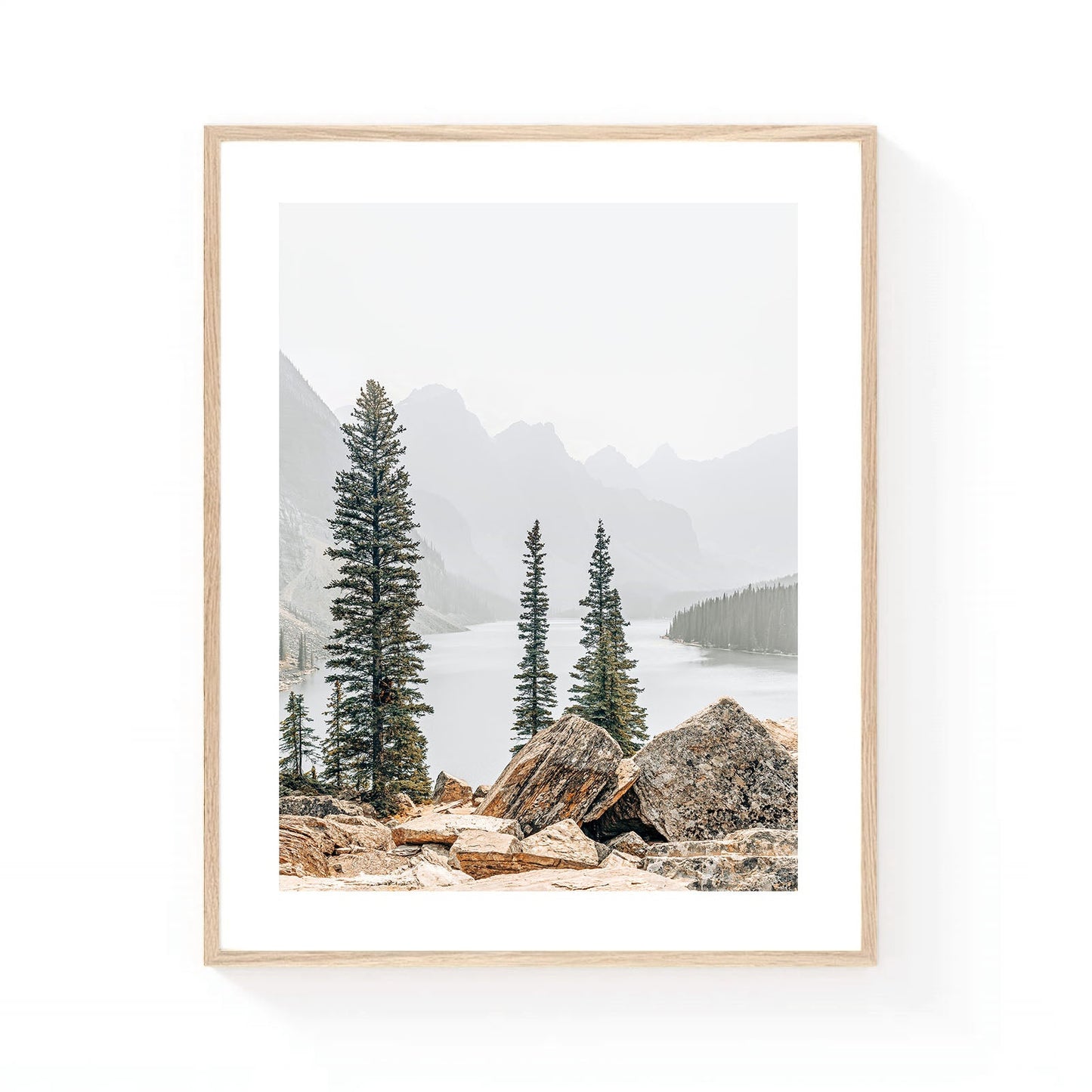 Mountain lake wall art featuring evergreen pine trees, rocky foreground boulders, misty alpine peaks, and calm water in a serene wilderness landscape.