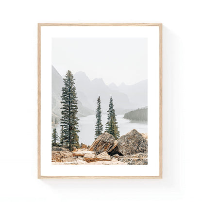 Mountain lake wall art featuring evergreen pine trees, rocky foreground boulders, misty alpine peaks, and calm water in a serene wilderness landscape.