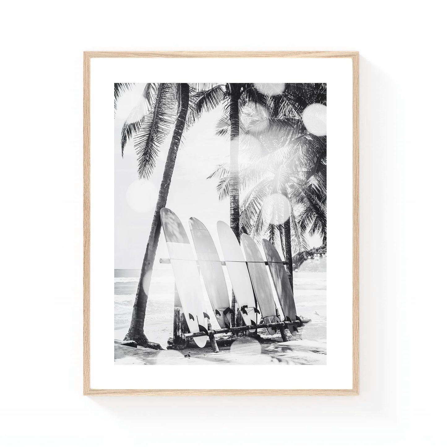 Black and white coastal wall art featuring a row of surfboards resting beneath tall palm trees on a tropical beach, with soft sunlight and ocean waves in the background.