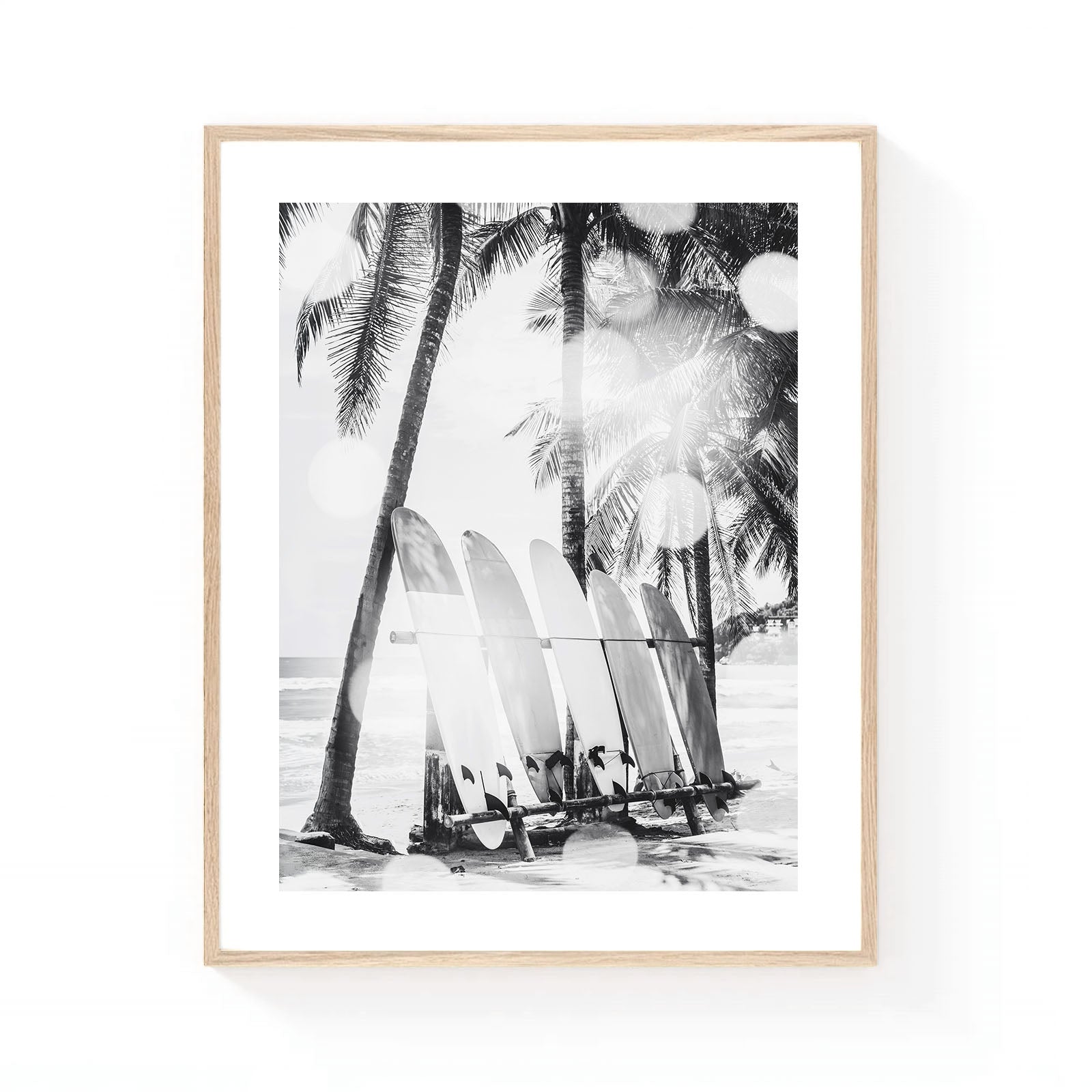 Black and white coastal wall art featuring a row of surfboards resting beneath tall palm trees on a tropical beach, with soft sunlight and ocean waves in the background.
