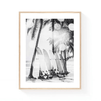 Black and white coastal wall art featuring a row of surfboards resting beneath tall palm trees on a tropical beach, with soft sunlight and ocean waves in the background.