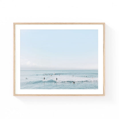 Surf wall art featuring surfers waiting beyond gentle ocean waves beneath a soft blue horizon in calming coastal tones.