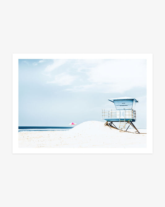 Huntington Beach wall art featuring Lifeguard Tower 14 and a colorful beach umbrella against bright sand and a soft blue coastal sky.