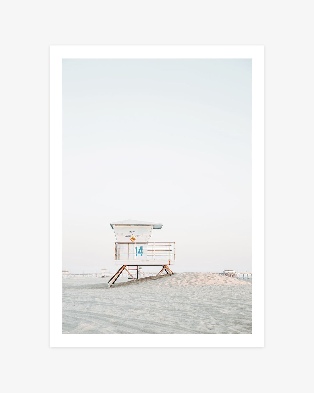 Huntington Beach lifeguard tower wall art featuring Tower 14 on a sandy rise beneath a pale coastal sky, with the pier in the distance.