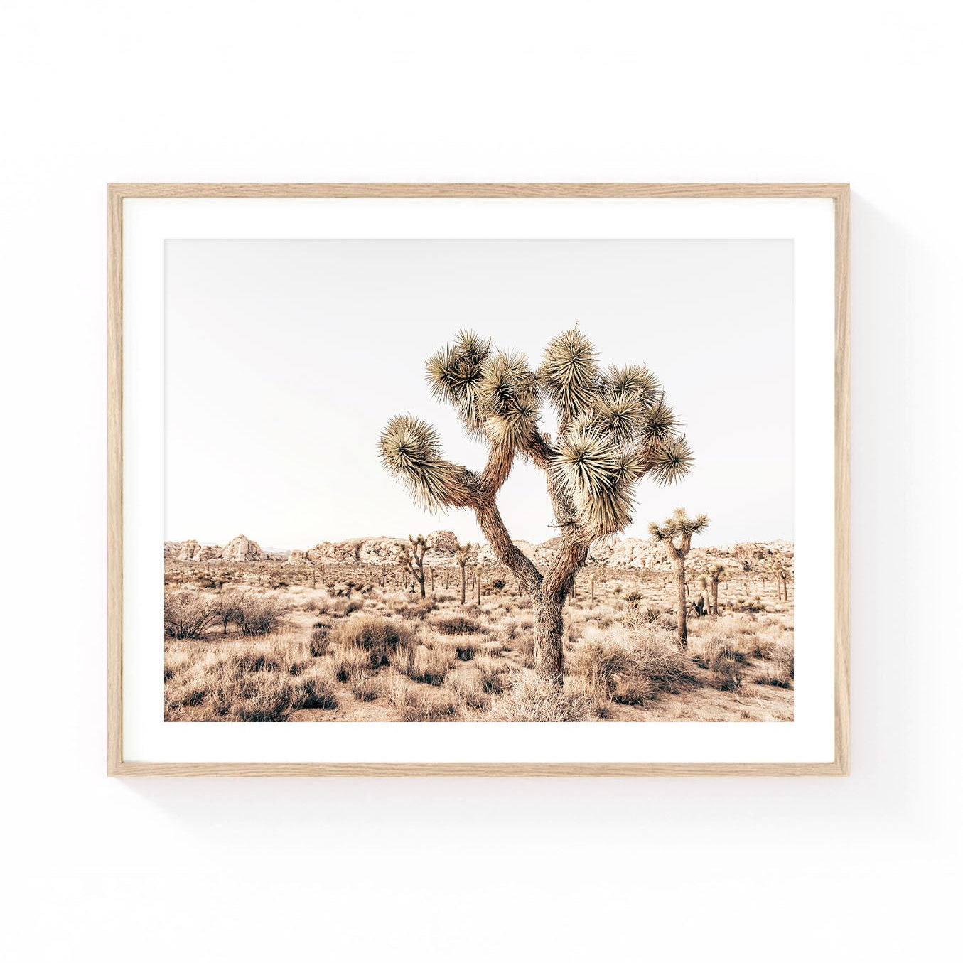 Joshua Tree wall art featuring a sculptural Joshua tree surrounded by desert grasses, rocky boulders, and expansive Mojave desert scenery in soft neutral tones.