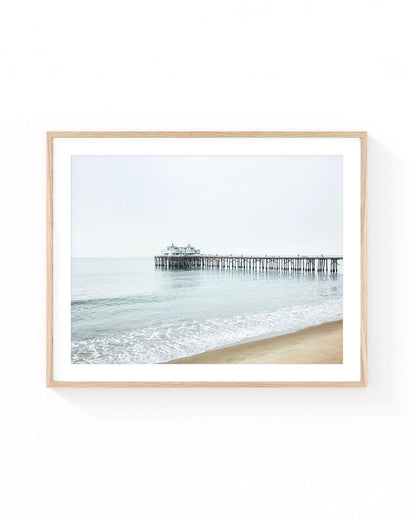 Malibu Pier wall art featuring the historic pier stretching over calm ocean water with gentle waves along a sandy beach under a soft overcast sky. Neutral coastal photography in light blue, beige, and white tones.