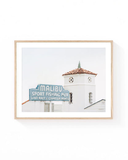 Malibu Pier wall art featuring the vintage Malibu Sport Fishing Pier sign beside a white coastal tower with a terracotta tile roof. Soft neutral California beach photography in muted blue, white, and warm terracotta tones.