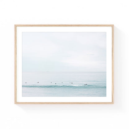 Minimalist surf wall art featuring surfers sitting in a calm ocean lineup beyond a soft rolling wave under a pale pastel sky.