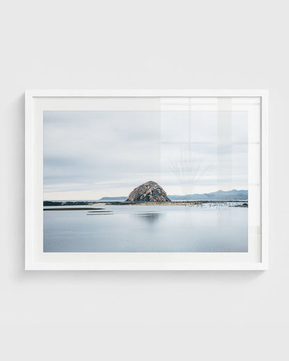 Morro Bay rock wall art featuring Morro Rock rising from calm reflective water with soft clouds and distant coastal hills under a muted sky.
