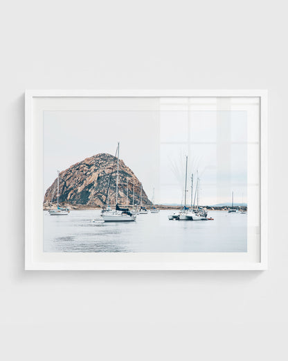 Morro Bay harbor with sailboats on calm water and Morro Rock rising in the background under a soft neutral sky.