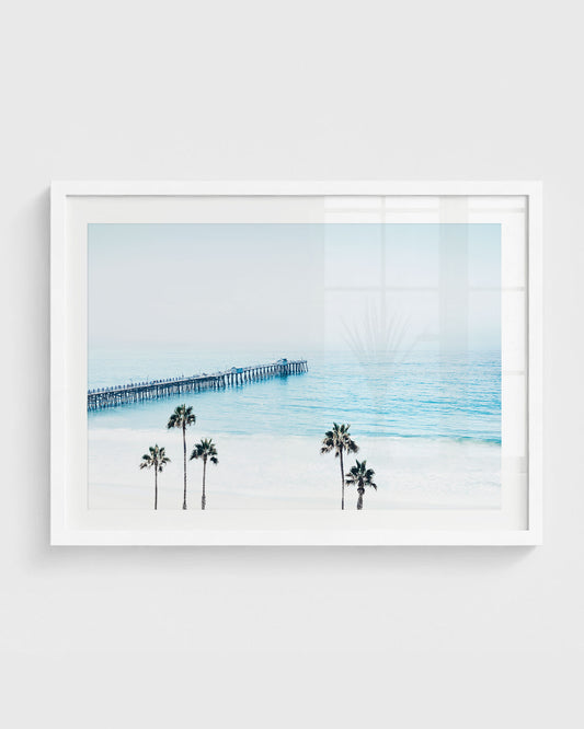 San Clemente Pier wall art featuring a long wooden pier over calm blue ocean with soft haze and palm trees in the foreground.