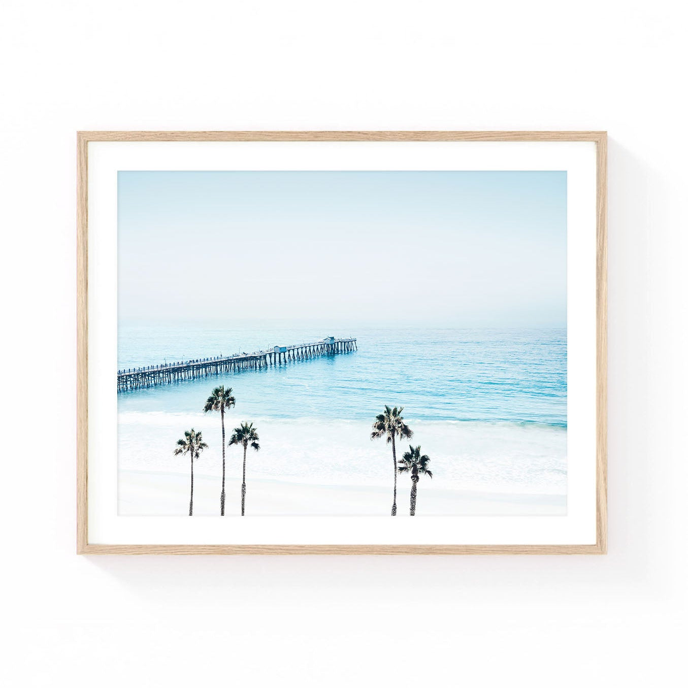 San Clemente Pier wall art featuring a long wooden pier extending into the Pacific Ocean, viewed from above with palm trees, soft blue water, white sand beach, and an airy coastal sky in light pastel tones.