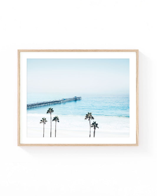San Clemente Pier wall art featuring a long wooden pier extending into the Pacific Ocean, viewed from above with palm trees, soft blue water, white sand beach, and an airy coastal sky in light pastel tones.
