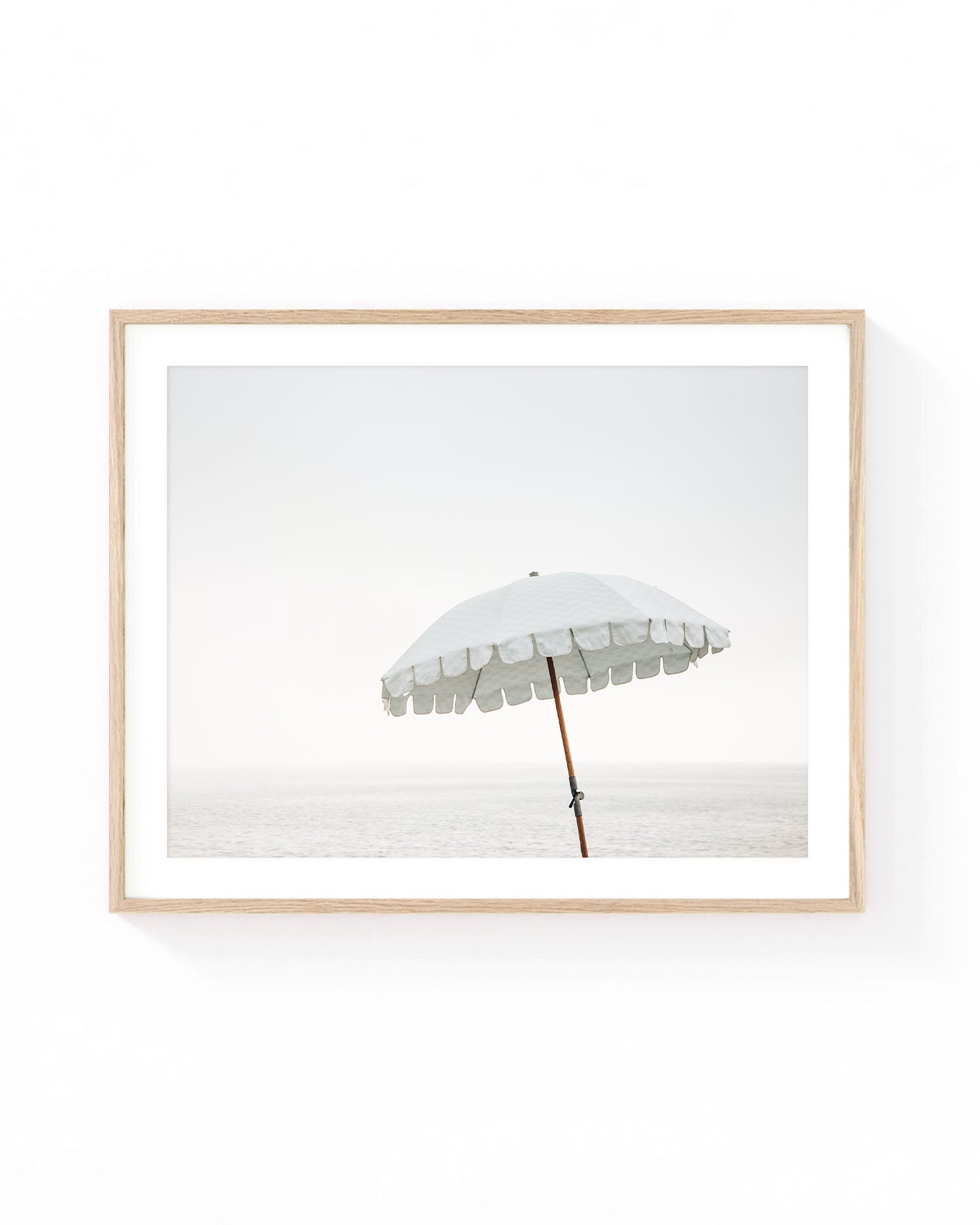 White beach umbrella wall art featuring a vintage-style parasol standing by the calm ocean under a soft hazy sky. Minimalist coastal photography in neutral white, beige, and pale blue tones.