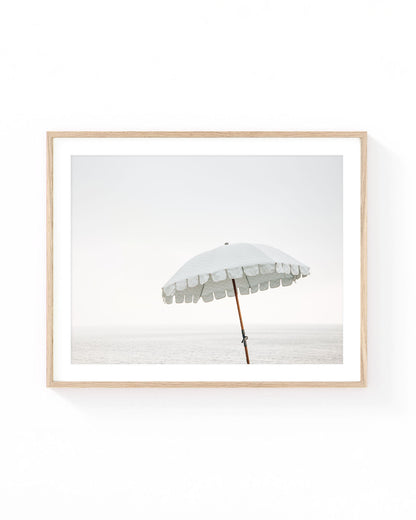 White beach umbrella wall art featuring a vintage-style parasol standing by the calm ocean under a soft hazy sky. Minimalist coastal photography in neutral white, beige, and pale blue tones.
